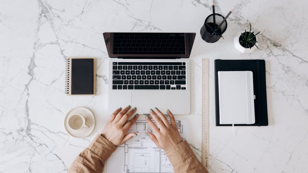 Top view of a stylish home office desk with a laptop, planner, and coffee cup, showing hands on a blueprint.